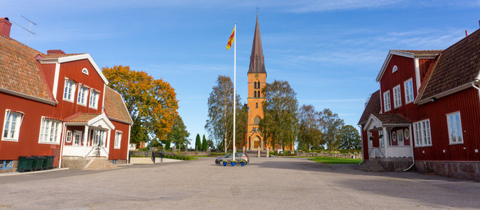 Hällestad kyrka i rött tegel syns i mitten på bilden. På båda sidor om kyrkan finns två röda trähus. I mitten finns också en flaggstång med Svenska kyrkans flagga.